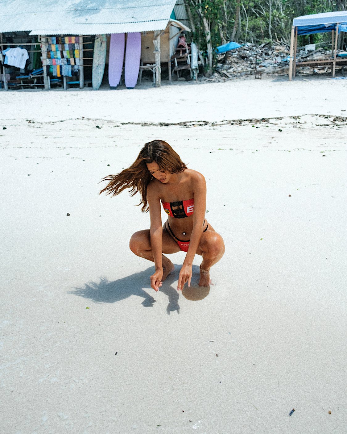 Front full body shot of the model sitting on the beach, highlighting the sexy Clip Nitro Bandeau bikini. The cute red bikini top is paired with high wasted, adjustable sides bikini bottoms.