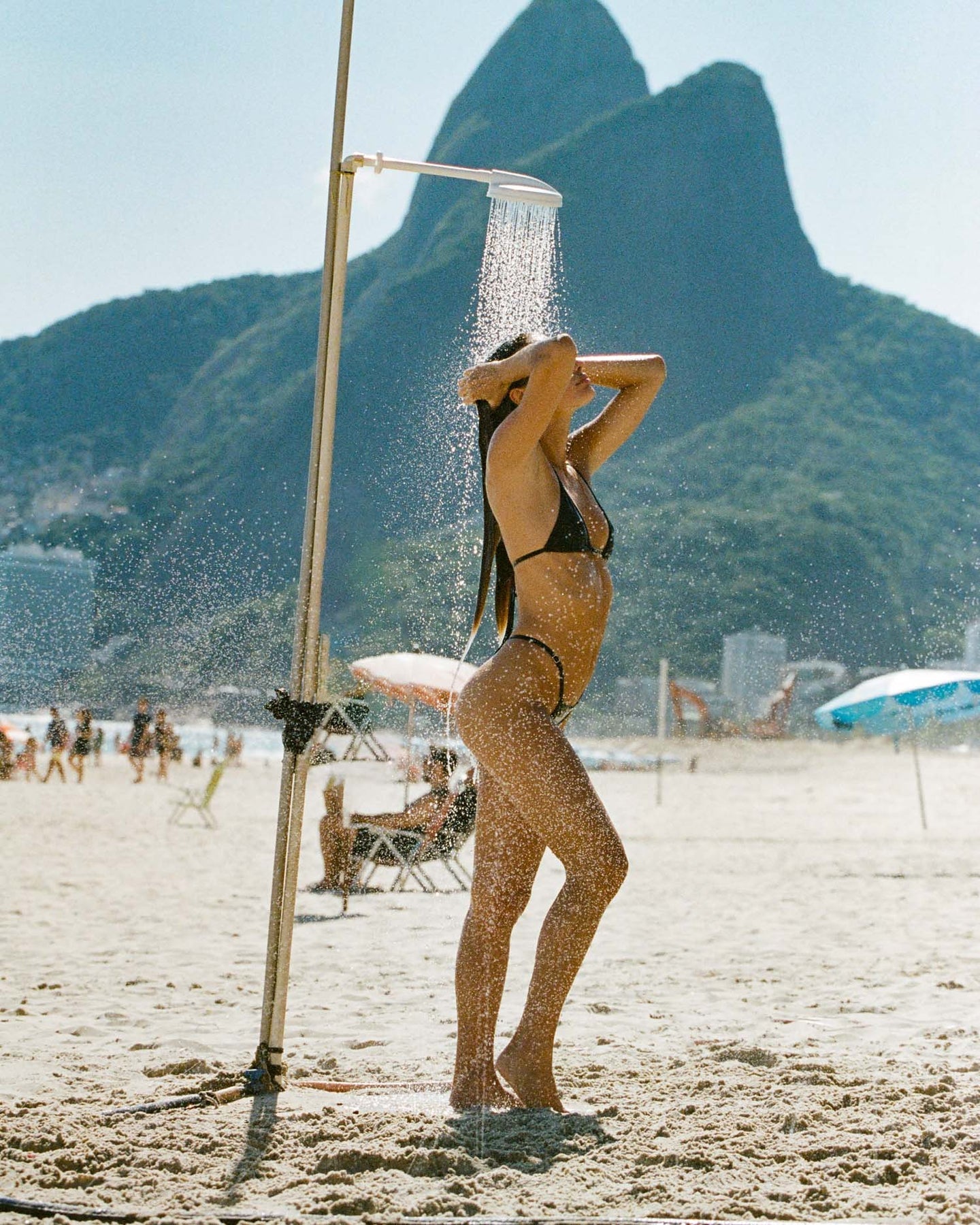 Woman in black bikini under beach shower with scenic mountain view in the background.