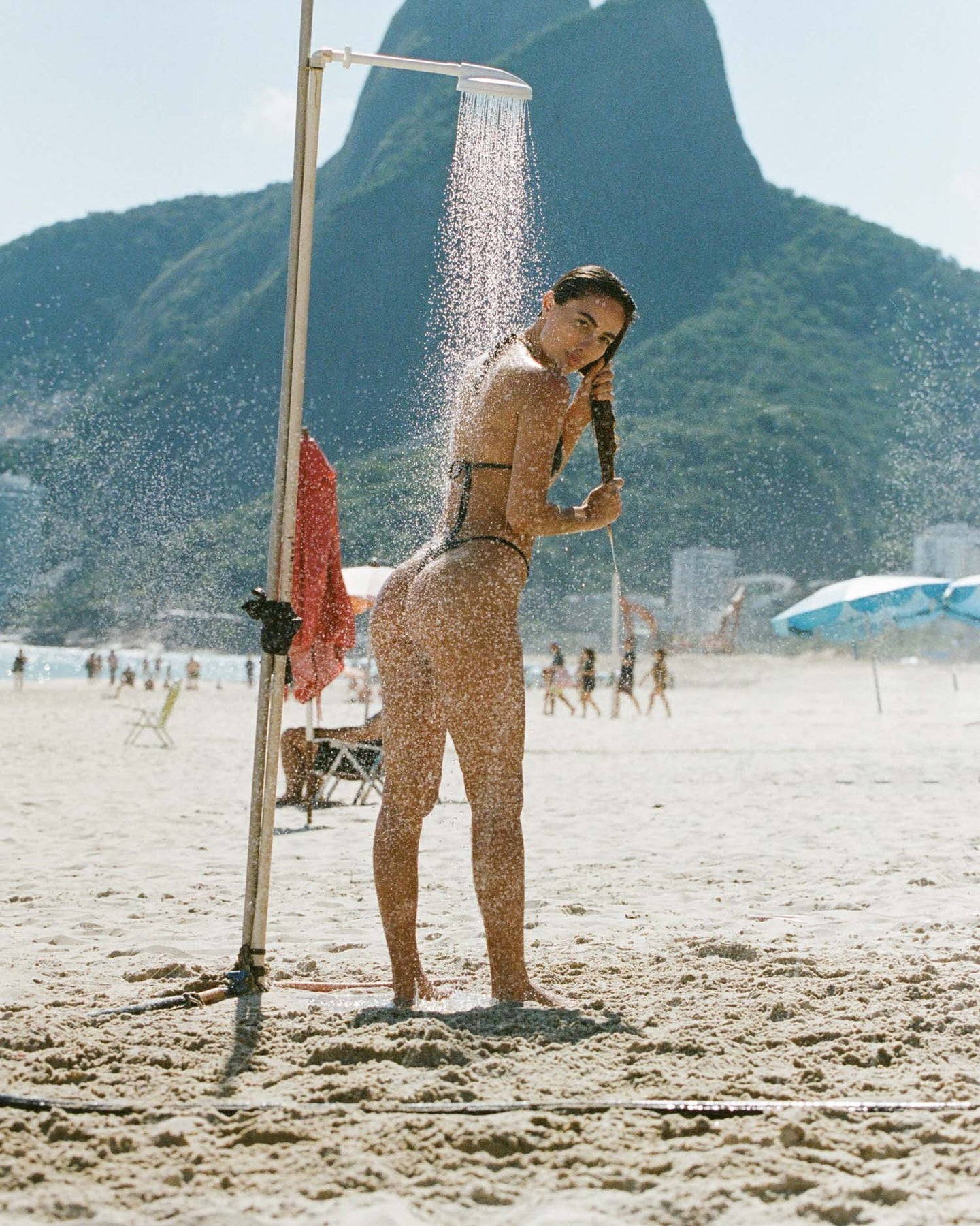 Woman in black bikini using outdoor beach shower with mountain view in the background.