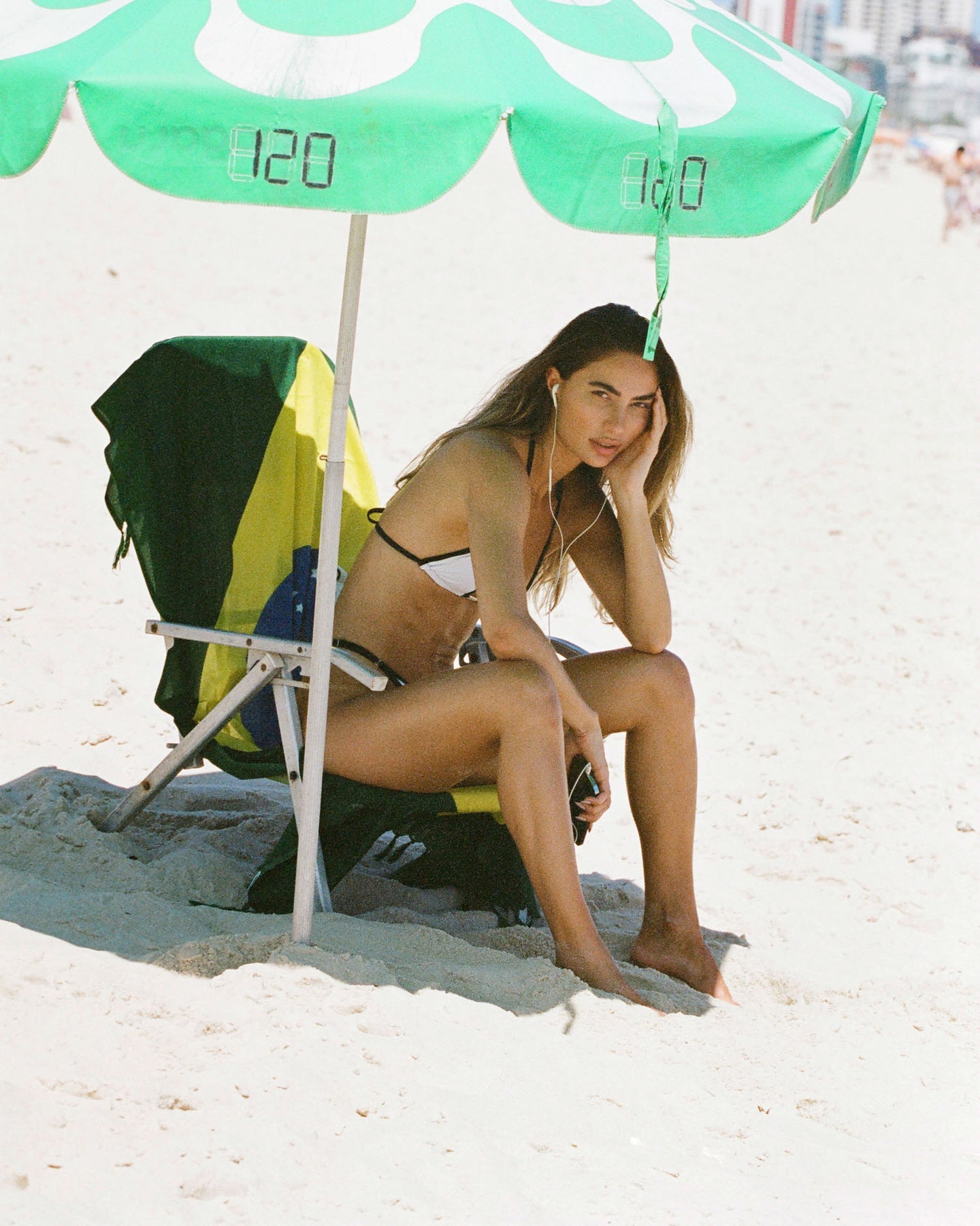 Seated gracefully and gazing into the distance, the model poses in comfortable swimwear featuring a serene white bikini with black outlines, adjustable side, and high-waisted cut.