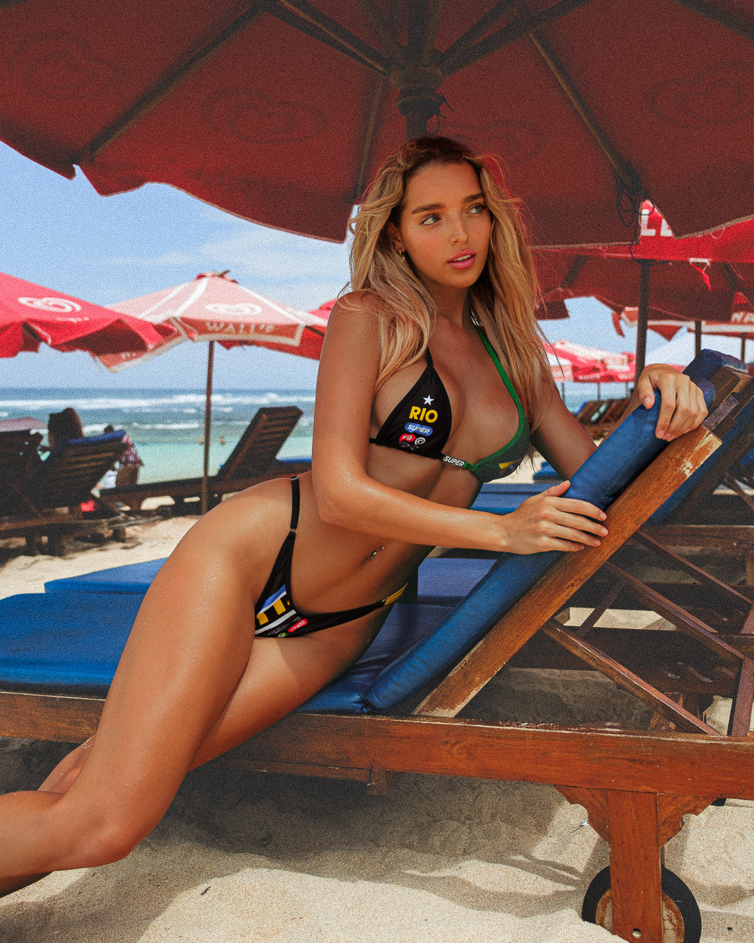 In a close-up side body shot, the model sits on a beach bench posing in a sexy, black women's bikini with Rio-inspired print. The classic triangle top is embellished with a cute charm bar, paired with high-waisted, adjustable-side thong bottoms, creating a cute and comfortable beach look.