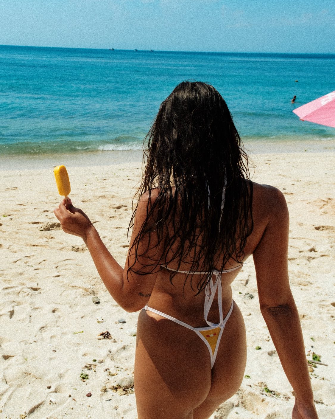 In a close-up rear view shot on the beach, the model showcases the high-waisted thong bottoms with adjustable sides in the vibrant yellow bikini. The white outlines add a stylish touch, creating a distinctive look for unique swimwear.