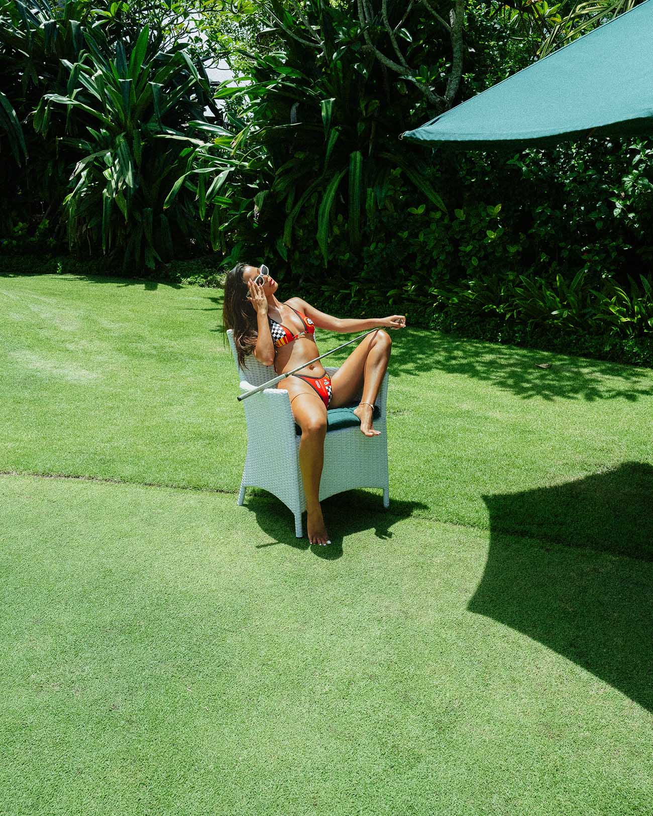 Model in Limit Red bikini relaxing on a chair in lush garden setting, showcasing sporty and vibrant swimwear style.