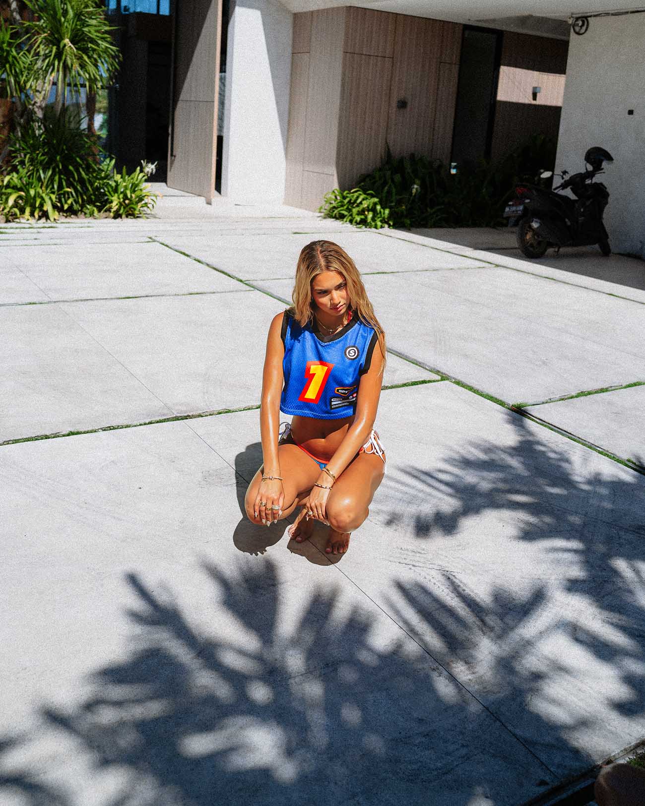 Model in a blue jersey crop top with sport graphics, crouching outdoors on a sunny day.