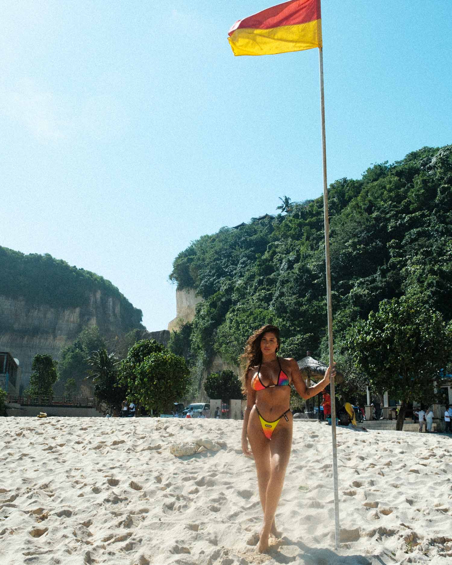 The beach becomes a runway as the model strikes a confident pose in the thong-style, high-waisted bikini with a captivating multihued gradient print. The distinctive triangle shape adds elegance to the scene, capturing the essence of a stylish summer beach day.