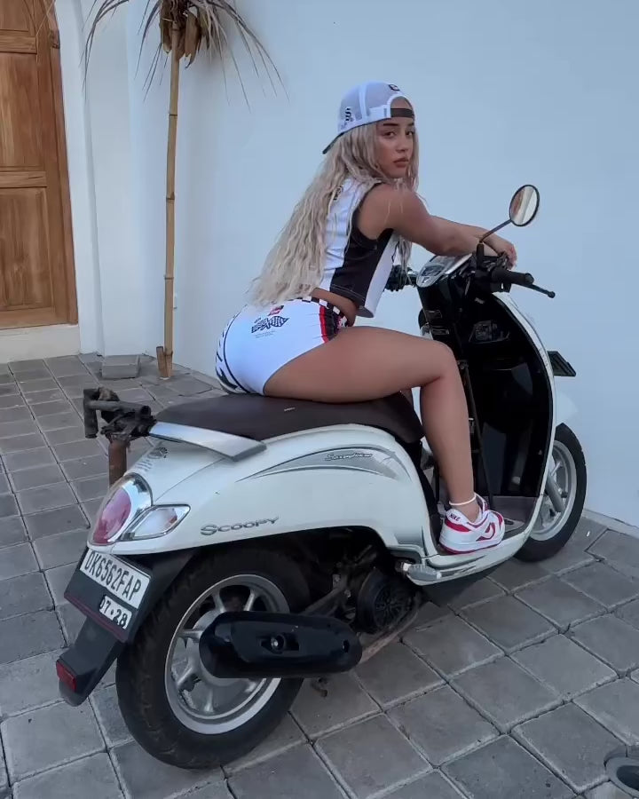 Model on a motorbike showcasing 360-degree view of trendy white short shorts with a checkered waistline and racing-inspired graphics.