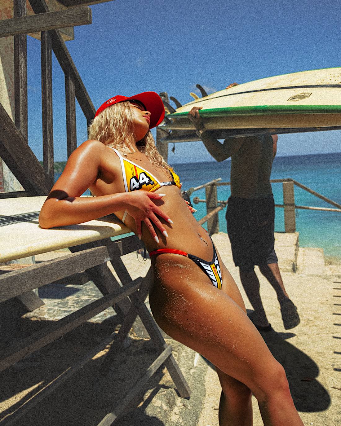 In a close-up side body shot, the model poses against a rail with an ocean backdrop, showcasing her stylish yellow women's bikini. The set features a triangle top embellished with a signature charm bar and high-waisted thong bottoms with adjustable sides. Complementing the look, she wears a red snapback for a cute, summery fit.