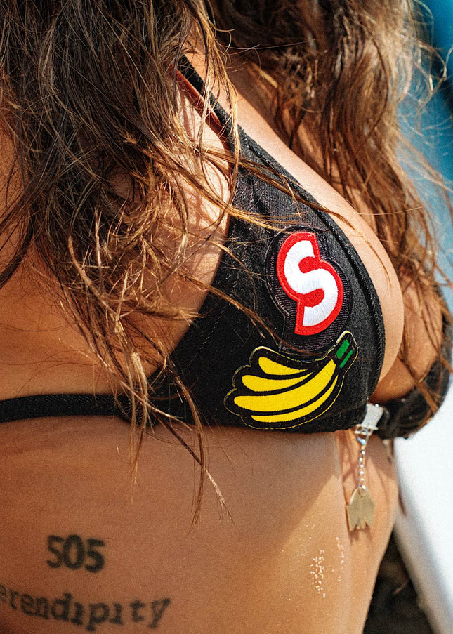 Extreme close-up detail of the woven patches on a high-waisted black denim bikini with tie sides. The intricate textures and patterns of the patches add a unique and stylish element to the overall design.