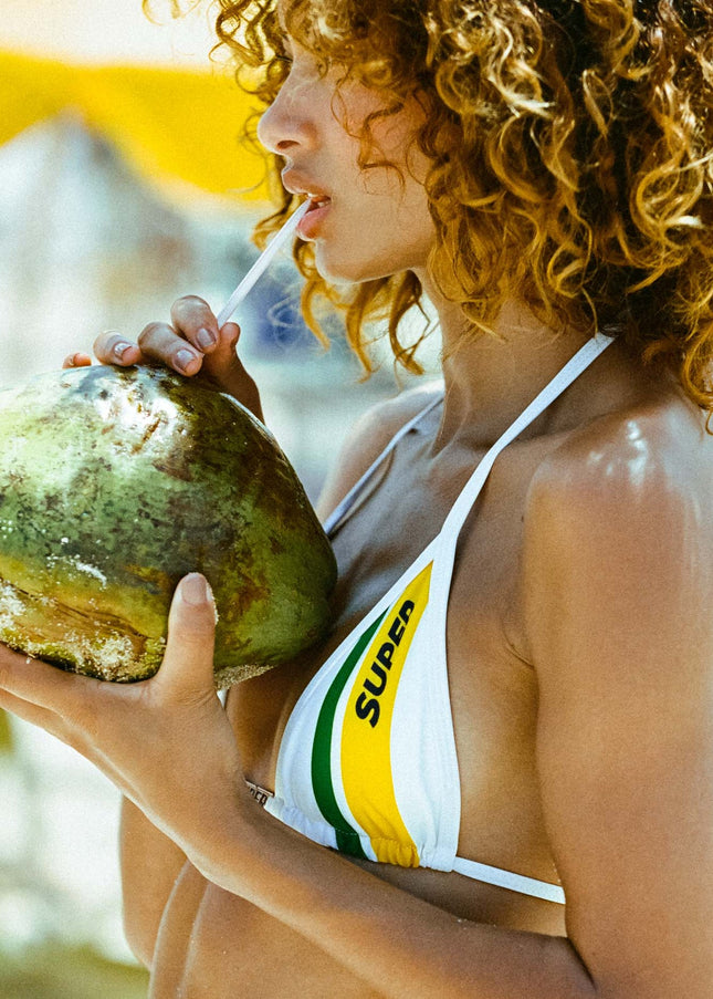 In a super close-up shot, the model enjoys a coconut while showcasing the details of the sexy white bikini's triangle top embellished with a charm bar and a Rio-inspired graphic.