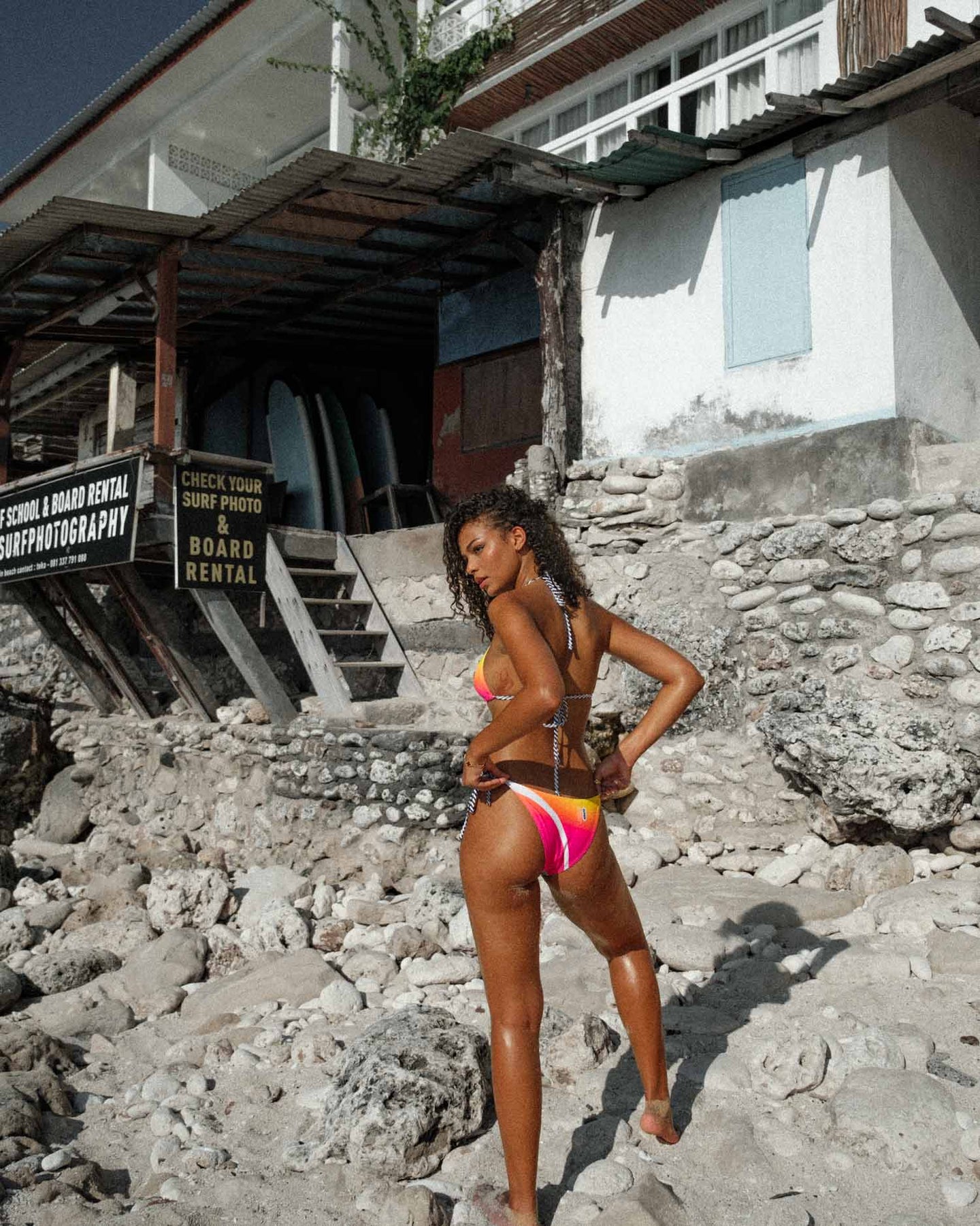 A rear full-body shot captures the model striking a confident pose in the Brazilian-style bikini with tie sides, adorned in a lively yellow, green, and pink print. The distinctive triangle shape and thong design create a bold silhouette against the backdrop of the beach, embodying the vibrant spirit of summer.