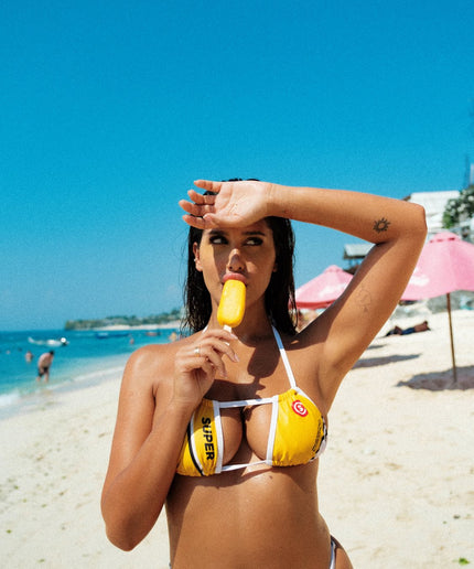 In a super close-up front shot on the beach, the model enjoys a popsicle while showcasing the vibrant yellow halterneck bikini top with crisp white outlines. This captivating look defines a sense of style for sexy bikini fashion.
