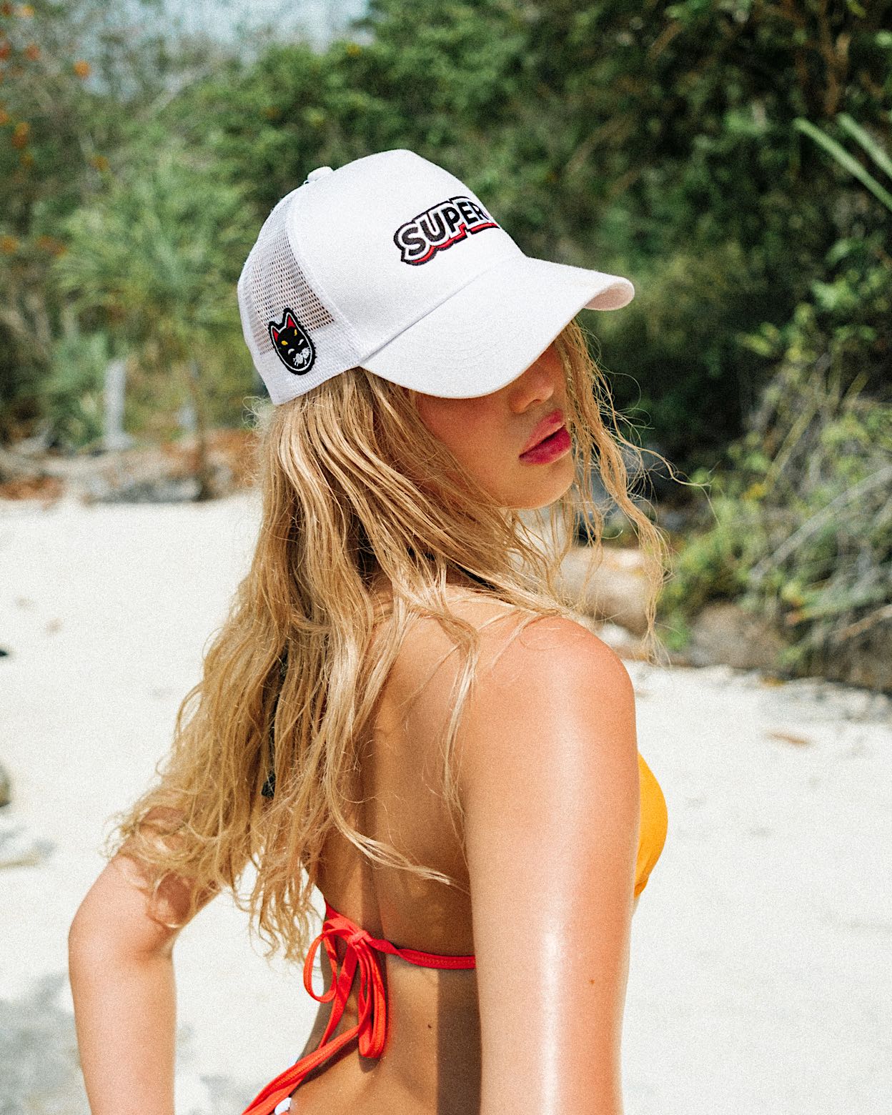 Close-up shot showcasing the top details, including the super white snapback hat, of a model in a High Waisted Palette Orange Thong bikini with adjustable sides and a vibrant pink, white, and black outline by the beach.