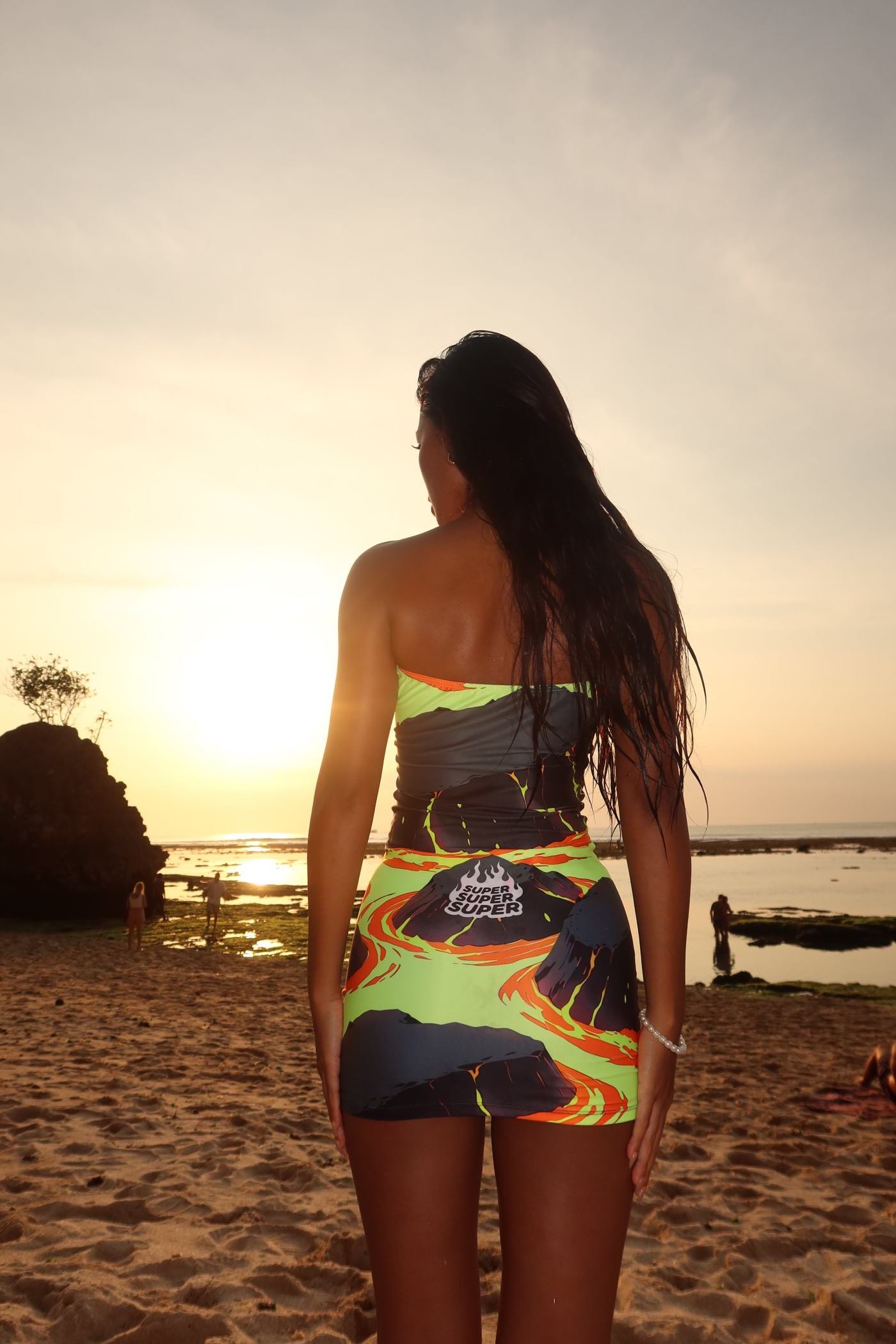 Woman in a vibrant Volcanic Tube Dress standing on a beach at sunset, showcasing the fiery anime design and molten magma motif on the back.